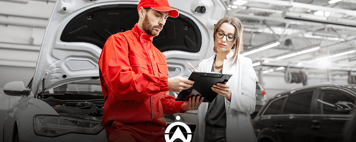 A mechanic in red overalls discusses vehicle details with a woman in a white blazer, indoors at an auto repair shop.