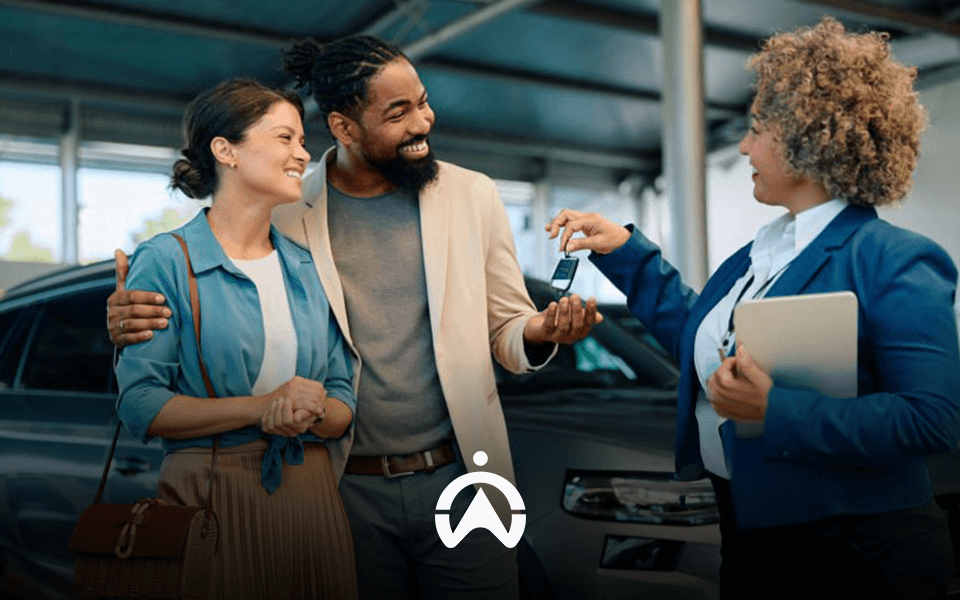 A couple happily interacts with a saleswoman at a car dealership, discussing keys in front of a parked vehicle.