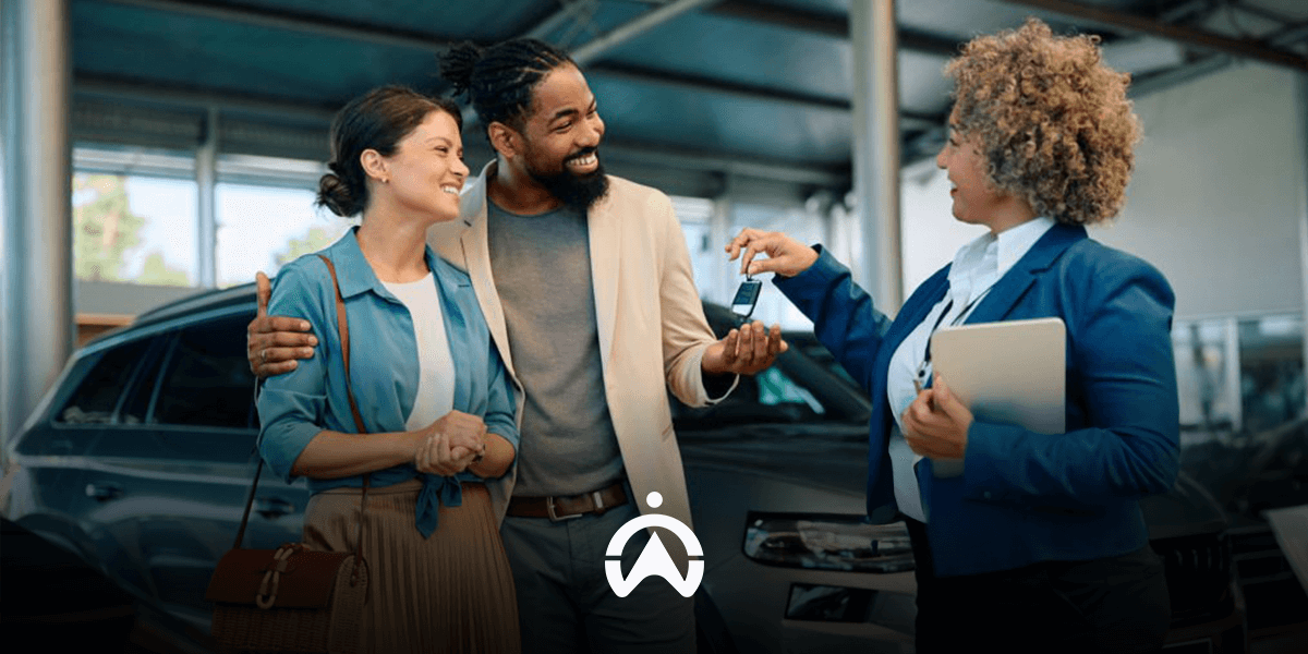 A couple happily interacts with a saleswoman at a car dealership, discussing keys in front of a parked vehicle.