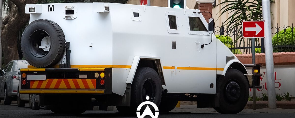 A white armored vehicle is stopped at a traffic light, with a right turn sign visible nearby in an urban setting.