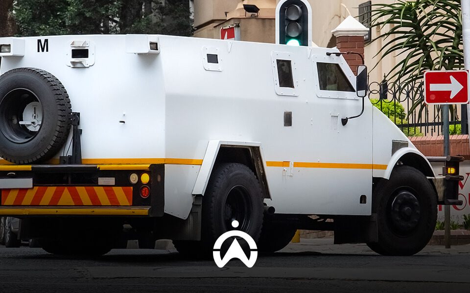A white armored vehicle is stopped at a traffic light, with a right turn sign visible nearby in an urban setting.