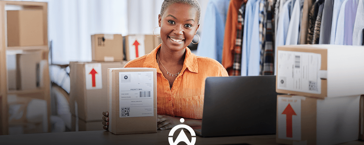 A person holds a labeled package in front of a laptop, surrounded by boxes, with clothes hanging in the background.