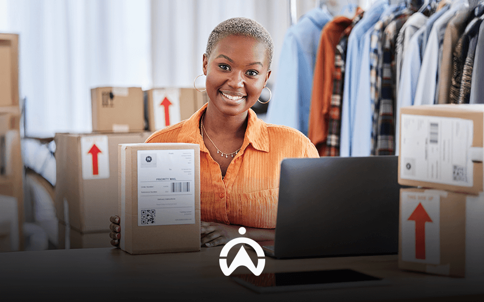 A person holds a labeled package in front of a laptop, surrounded by boxes, with clothes hanging in the background.