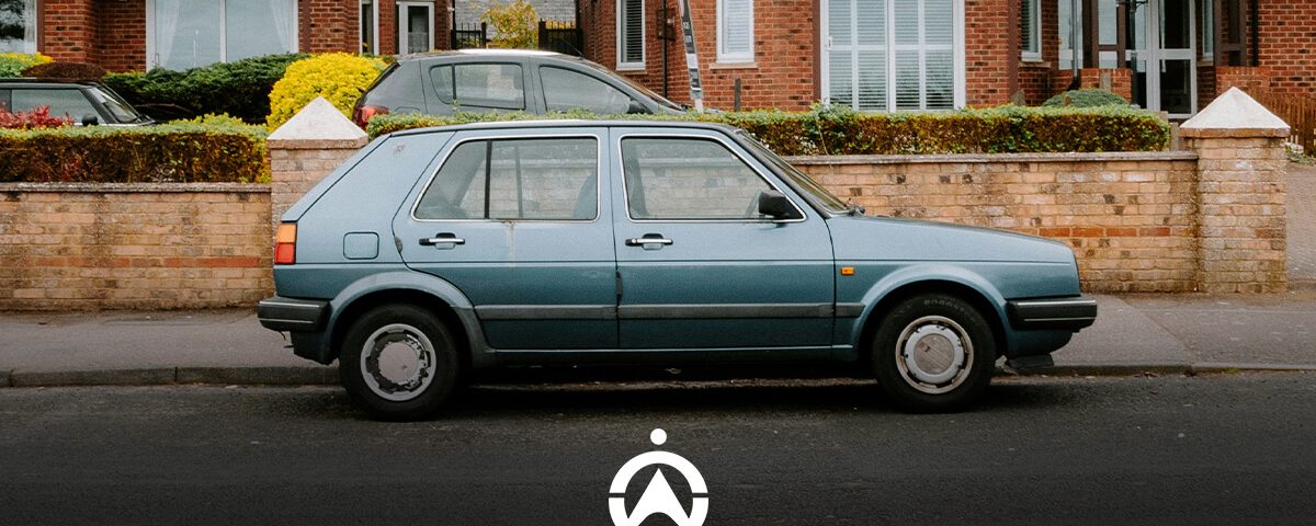 A vintage blue hatchback parked on a residential street beside hedges, with brick houses in the background and a clear sky above.