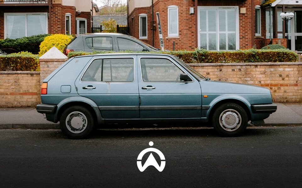 A vintage blue hatchback parked on a residential street beside hedges, with brick houses in the background and a clear sky above.