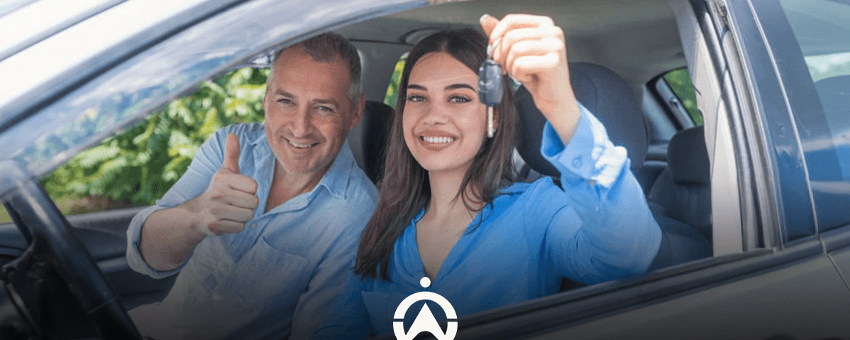 A woman in a blue shirt holds car keys while sitting in a vehicle, smiling with a man giving a thumbs up beside her.