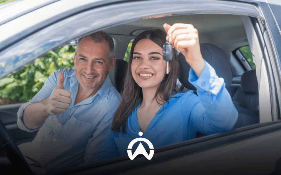 A woman in a blue shirt holds car keys while sitting in a vehicle, smiling with a man giving a thumbs up beside her.