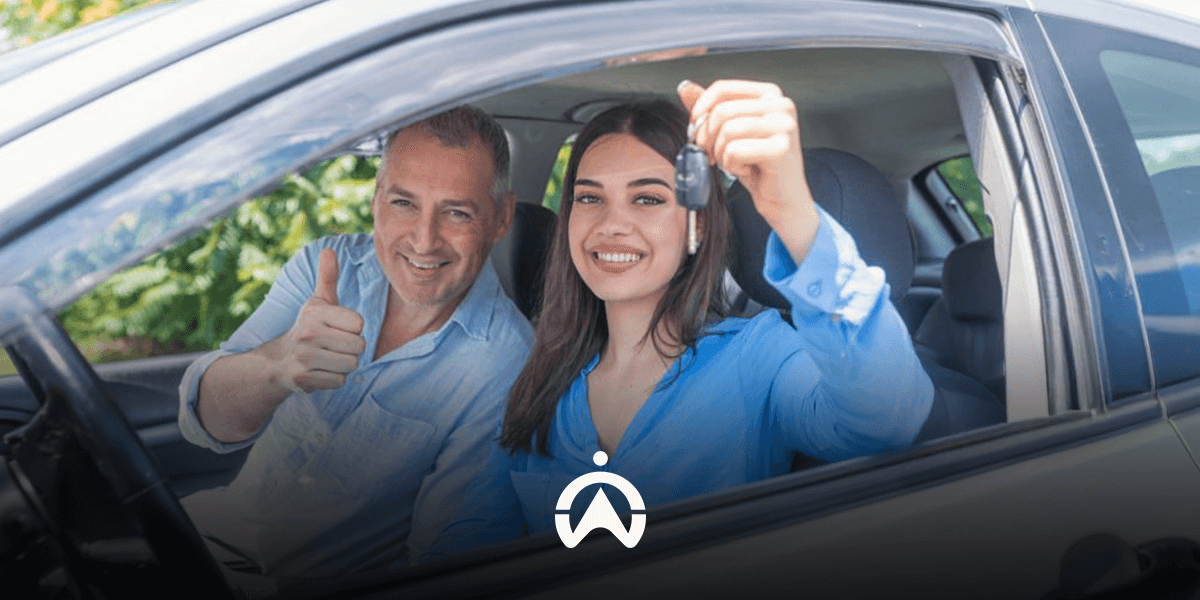 A woman in a blue shirt holds car keys while sitting in a vehicle, smiling with a man giving a thumbs up beside her.