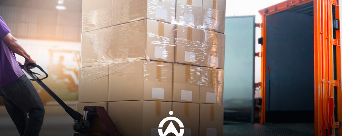 A worker uses a hand truck to transport a stack of wrapped boxes towards an open delivery truck in a warehouse setting.