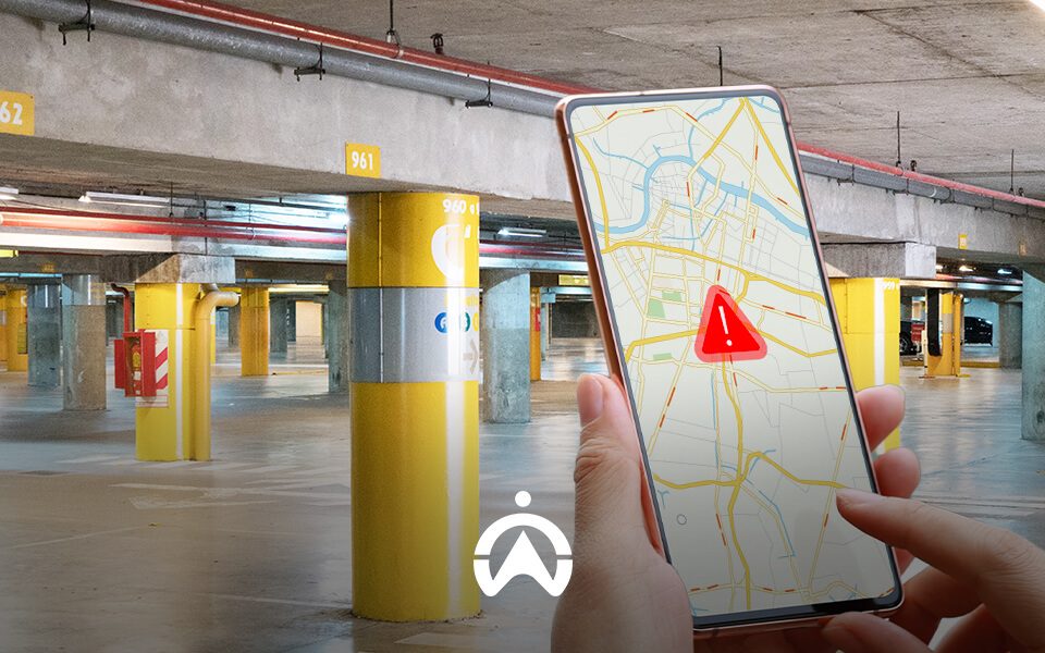 A person holds a smartphone displaying a map with an alert icon, surrounded by a dimly lit parking garage with numbered pillars.