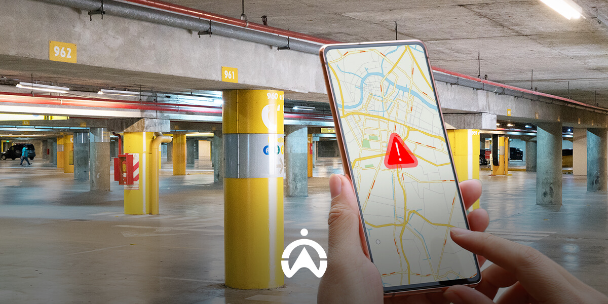 A person holds a smartphone displaying a map with an alert icon, surrounded by a dimly lit parking garage with numbered pillars.