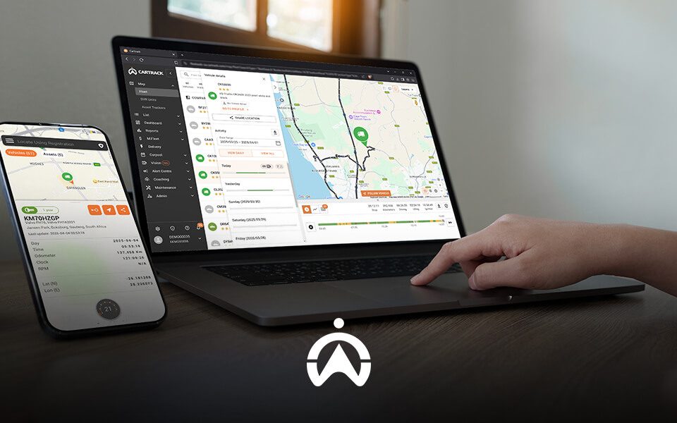 A person uses a laptop and smartphone displaying a vehicle tracking application, with a map and vehicle details visible on the screen.
