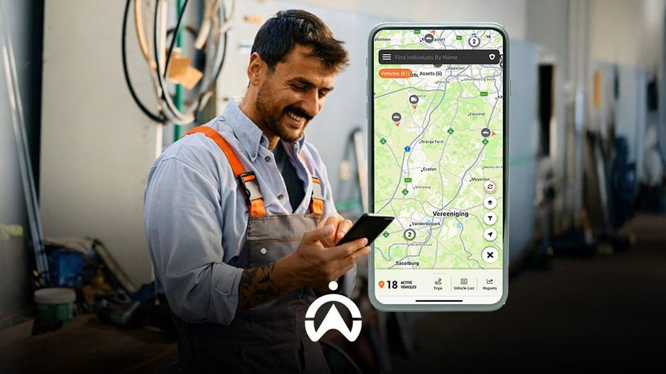 A worker in a workshop checks a smartphone displaying a map with vehicle and asset tracking information. Tools and equipment are visible in background.