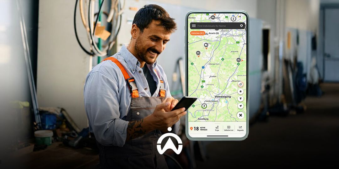A worker in a workshop checks a smartphone displaying a map with vehicle and asset tracking information. Tools and equipment are visible in background.