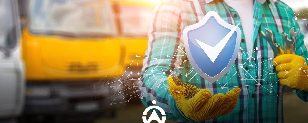 Worker in a plaid shirt and gloves holds a shield icon symbolizing safety, with yellow trucks blurred in the background.