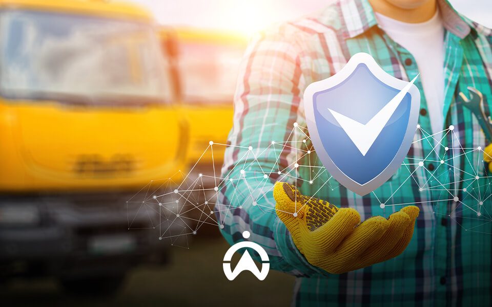 Worker in a plaid shirt and gloves holds a shield icon symbolizing safety, with yellow trucks blurred in the background.