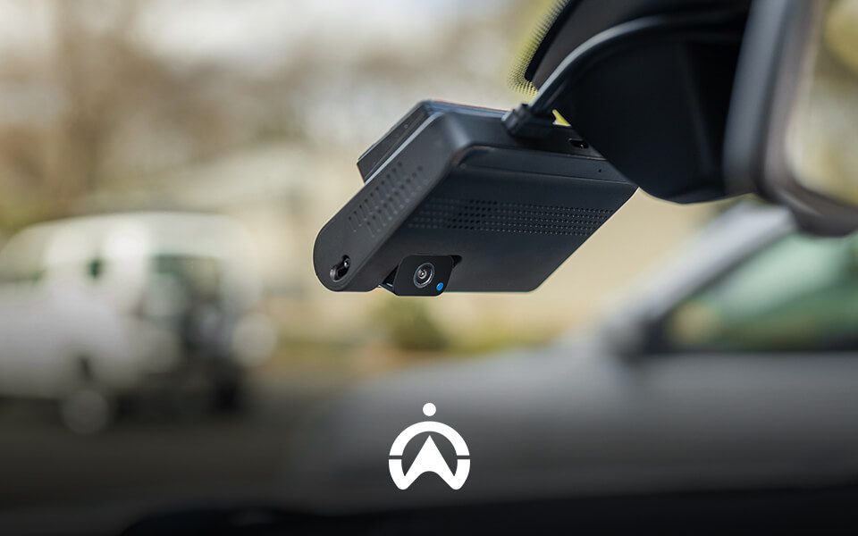 A close-up of a car dash cam mounted on a windshield, with a blurred white vehicle visible in the background.