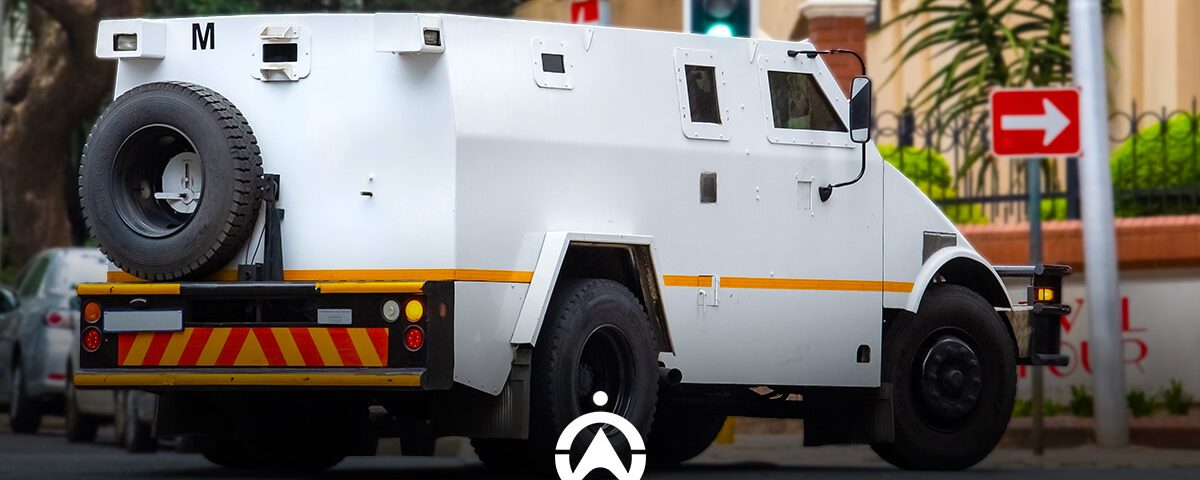 A white armored vehicle with yellow stripes parked on a street, displaying a distinctive wheel mounted on the rear. Traffic lights visible.