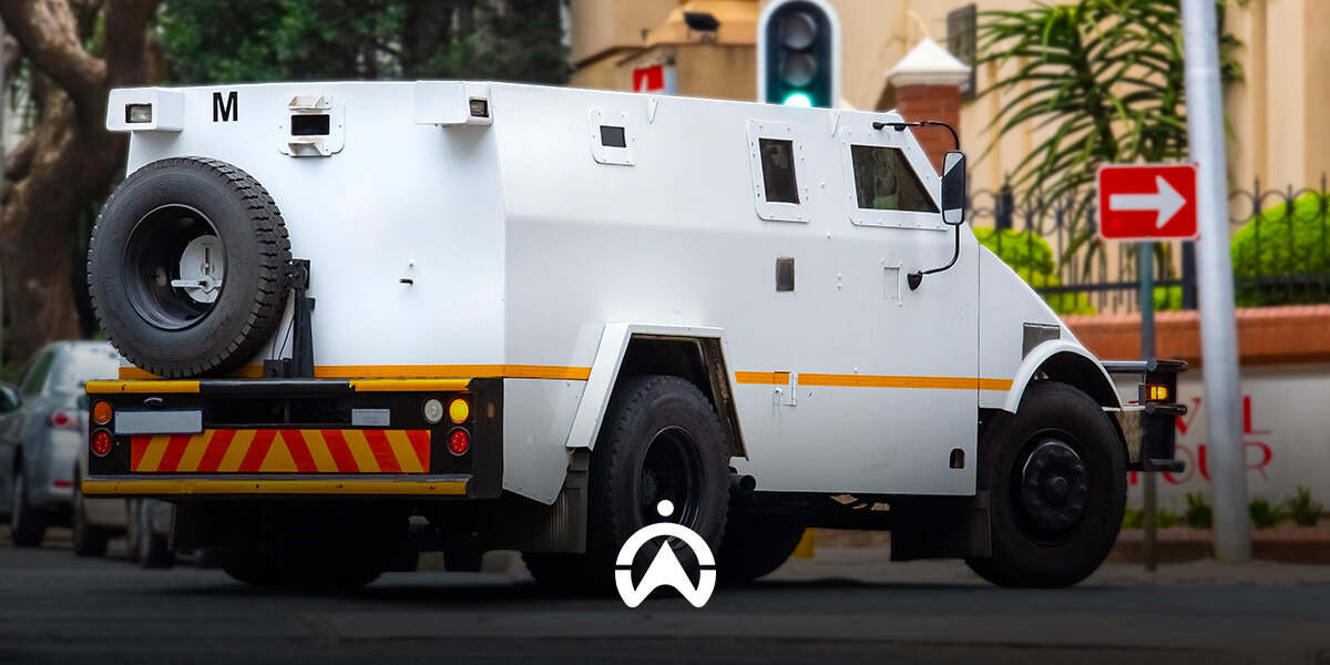 A white armored vehicle with yellow stripes parked on a street, displaying a distinctive wheel mounted on the rear. Traffic lights visible.