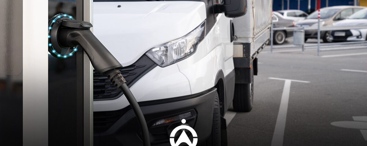 An electric vehicle charging cable inserted into a station, with a white delivery truck parked in the background.