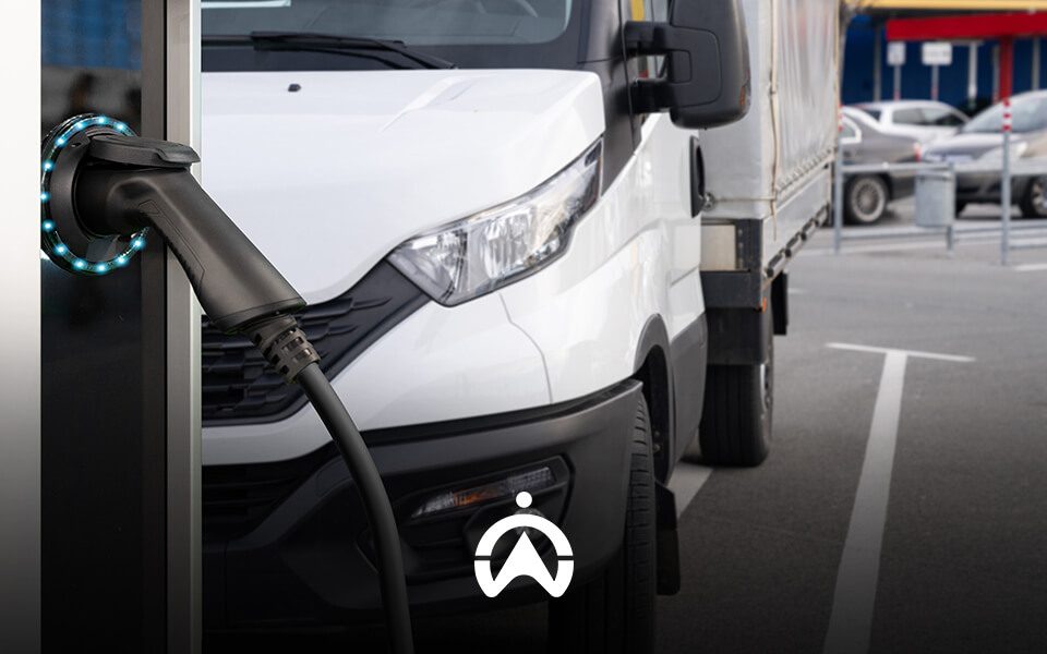 An electric vehicle charging cable inserted into a station, with a white delivery truck parked in the background.