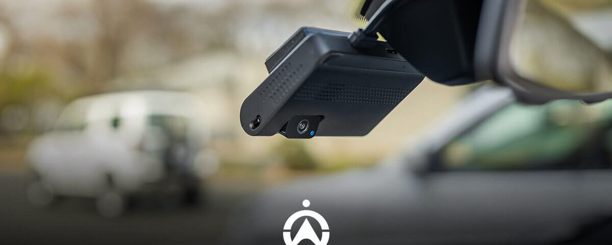 A compact dashboard camera mounted near a car's rearview mirror, capturing road views with blurred vehicles in the background.