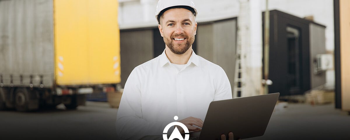 A construction worker in a white shirt and hard hat stands holding a laptop in a warehouse with shipping containers in the background.