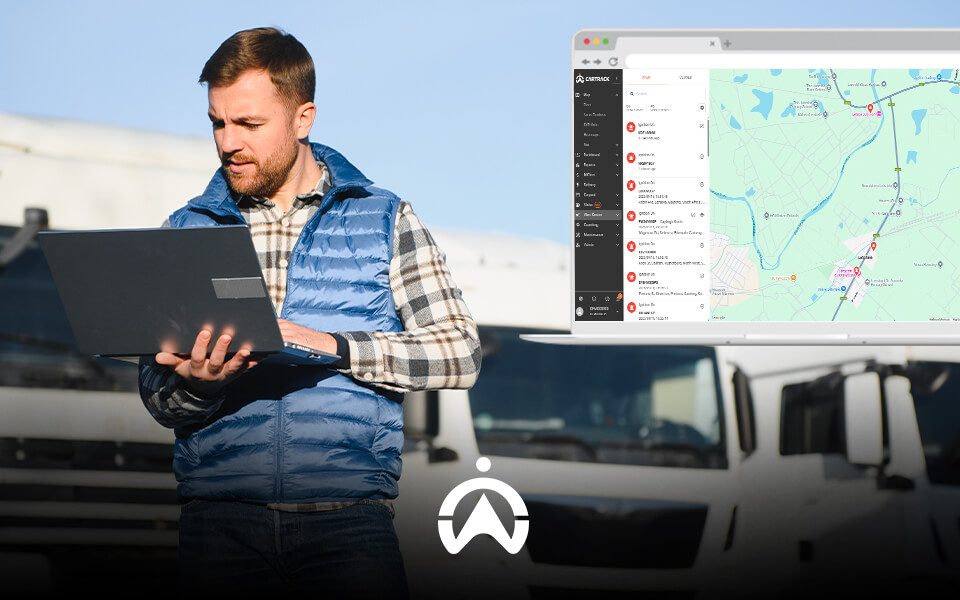 A man in a blue vest stands with a laptop, analyzing a map on the screen and surrounded by trucks in a parking area.