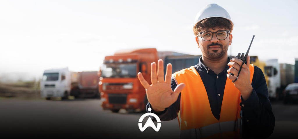 A construction worker in an orange vest and hard hat signals to stop while holding a walkie-talkie, with trucks in the background.