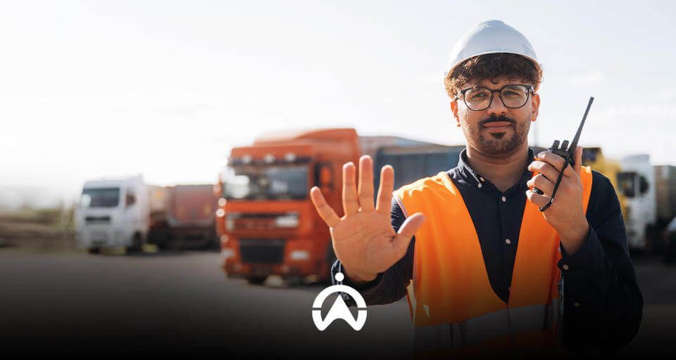 A construction worker in an orange vest and hard hat signals to stop while holding a walkie-talkie, with trucks in the background.