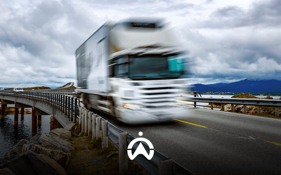A blurred white truck speeds over a curved bridge by a body of water under a dramatic sky, symbolizing motion and transport.