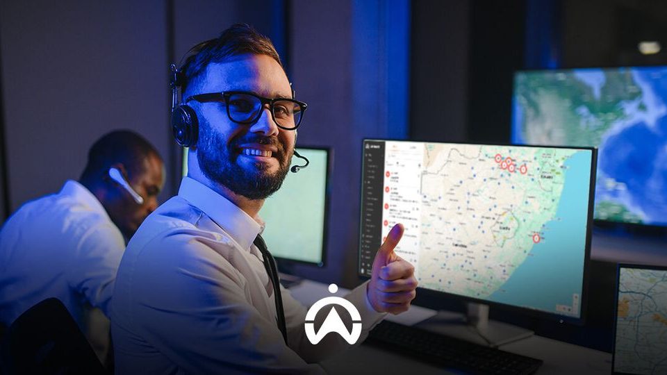 A person in a headset gives a thumbs up while operating multiple computer screens displaying maps and data in a control room.