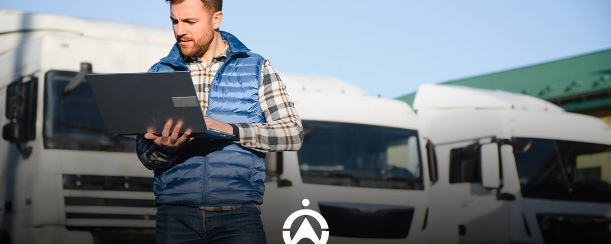 A man in a blue vest stands outside with a laptop, surrounded by parked white trucks under a clear blue sky.