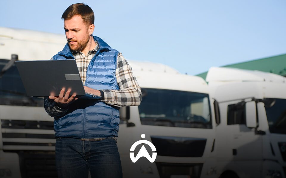 A man in a blue vest stands outside with a laptop, surrounded by parked white trucks under a clear blue sky.
