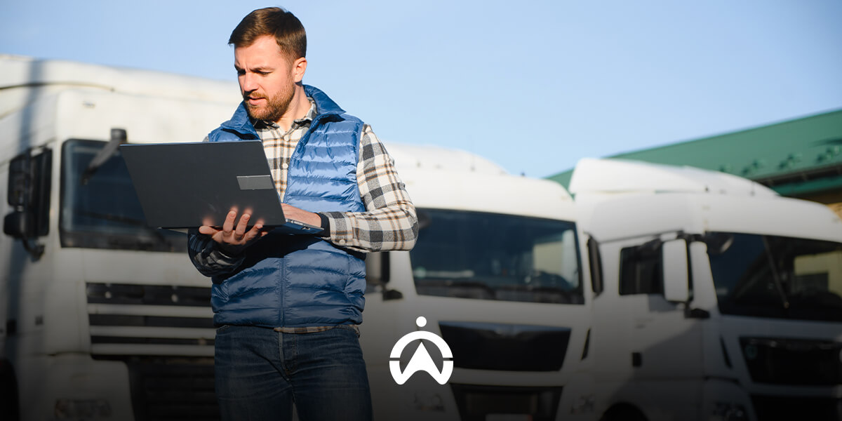 A man in a blue vest stands outside with a laptop, surrounded by parked white trucks under a clear blue sky.