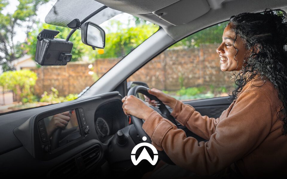 A person with curly hair drives a car, focused on the road ahead, with a dashboard camera mounted on the windshield.