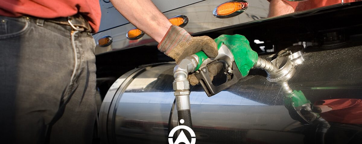 A person wearing gloves refuels a large truck, holding a green fuel nozzle against the vehicle's tank.
