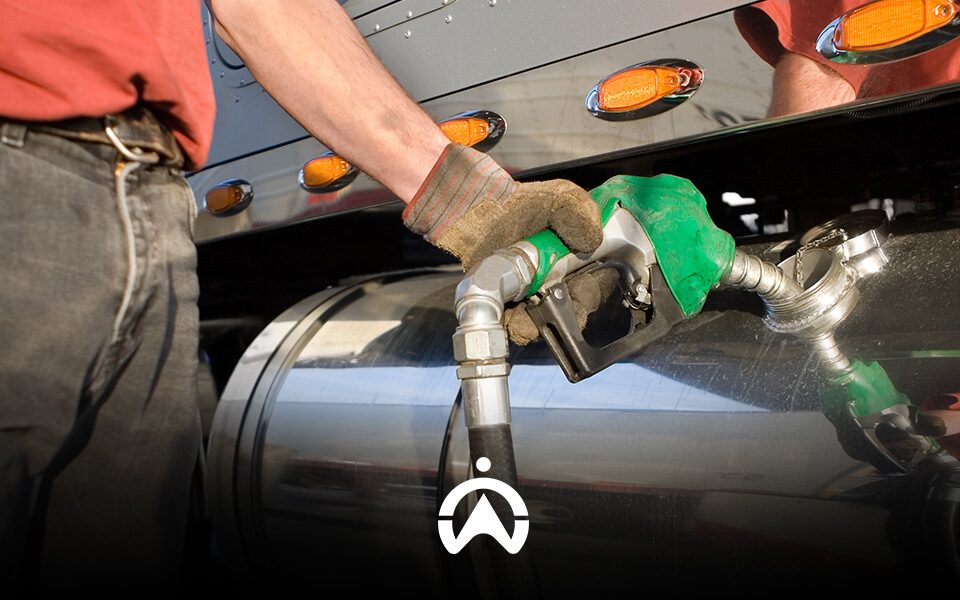 A person wearing gloves refuels a large truck, holding a green fuel nozzle against the vehicle's tank.