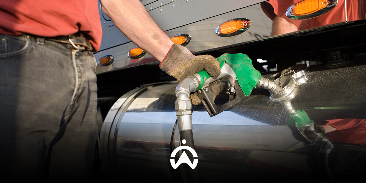 A person wearing gloves refuels a large truck, holding a green fuel nozzle against the vehicle's tank.